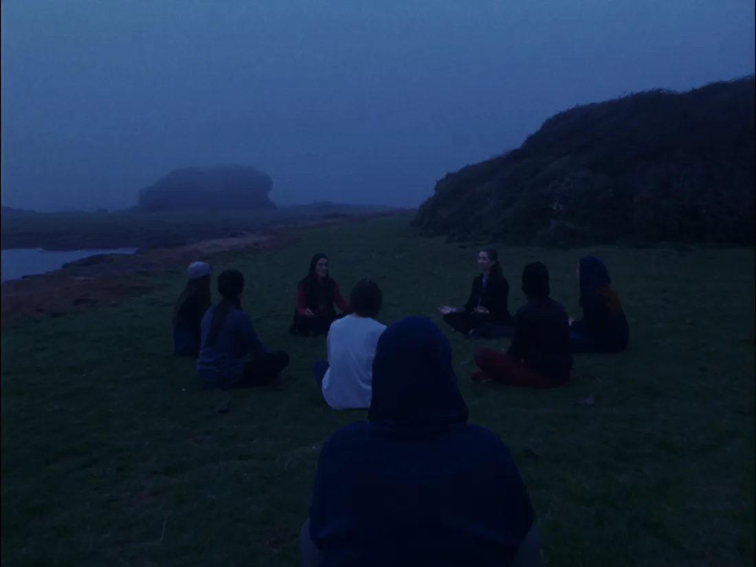 A circle of people seated on grass at blue hour, breathing together before the plunge